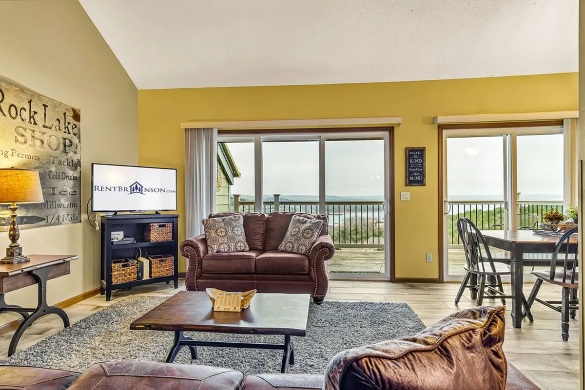 A spacious, open-concept interior featuring a cozy leather sofa facing large windows, with lake views just beyond. The room flows seamlessly into a dining area, emphasizing the open layout ideal for group gatherings. Decorative wall art and plenty of natural light contribute to an inviting and comfortable atmosphere.