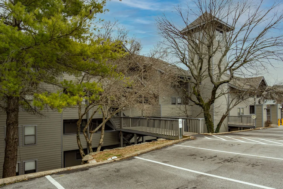 An inviting exterior scene featuring a wooden walkway winding through leafy trees, with a distinctive tower structure visible in the background. The setting feels tranquil and seamlessly blends the built environment with nature, suggesting easy access to outdoor activities.