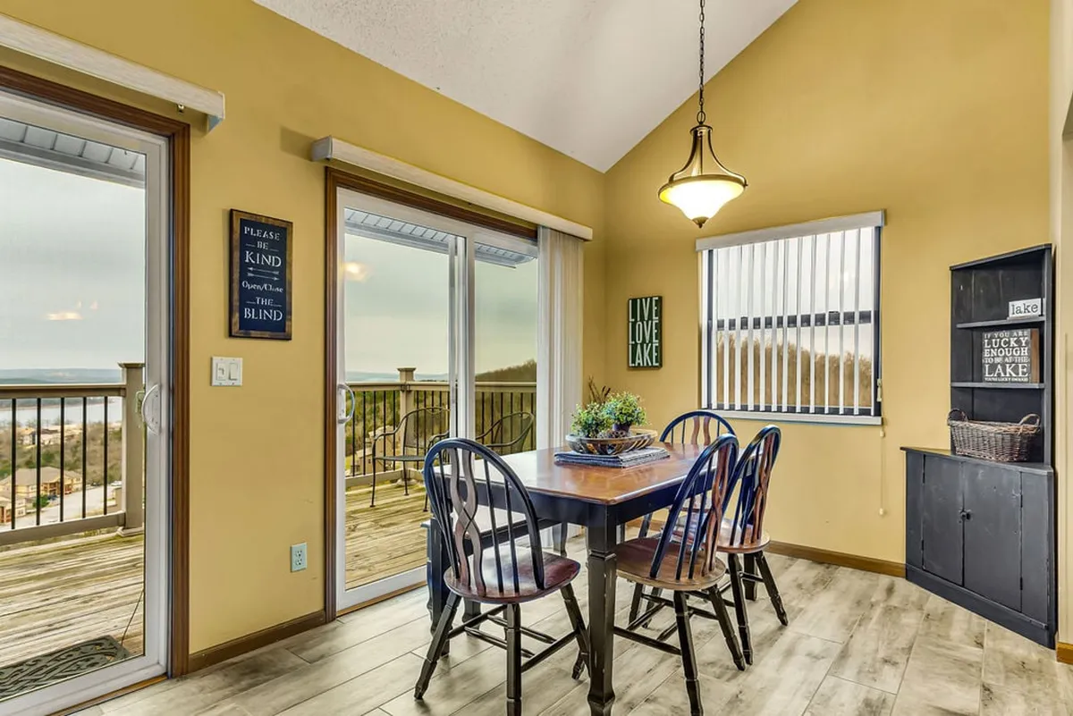 A cozy dining area centered around a large wooden table, framed by expansive windows. Decorative wall signs and a hanging light fixture create a welcoming atmosphere, while the lake can be glimpsed through the windows.