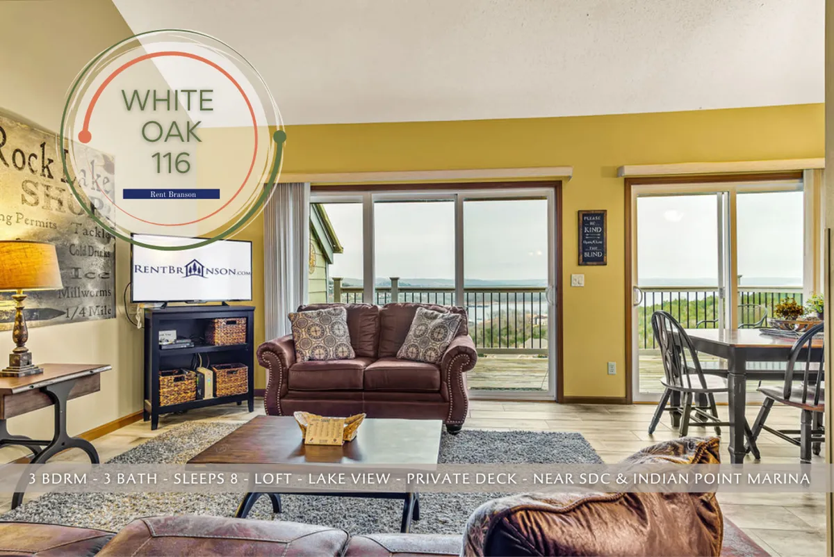 A welcoming living room with a comfortable seating area arranged around a central table, large windows flooding the room with natural light, and a decorative wall with uplifting text. Beyond the windows, a beautiful view of Table Rock Lake is visible, emphasizing the connection between indoor comfort and nature. The arrangement encourages socializing, relaxation, and enjoying the scenic backdrop.