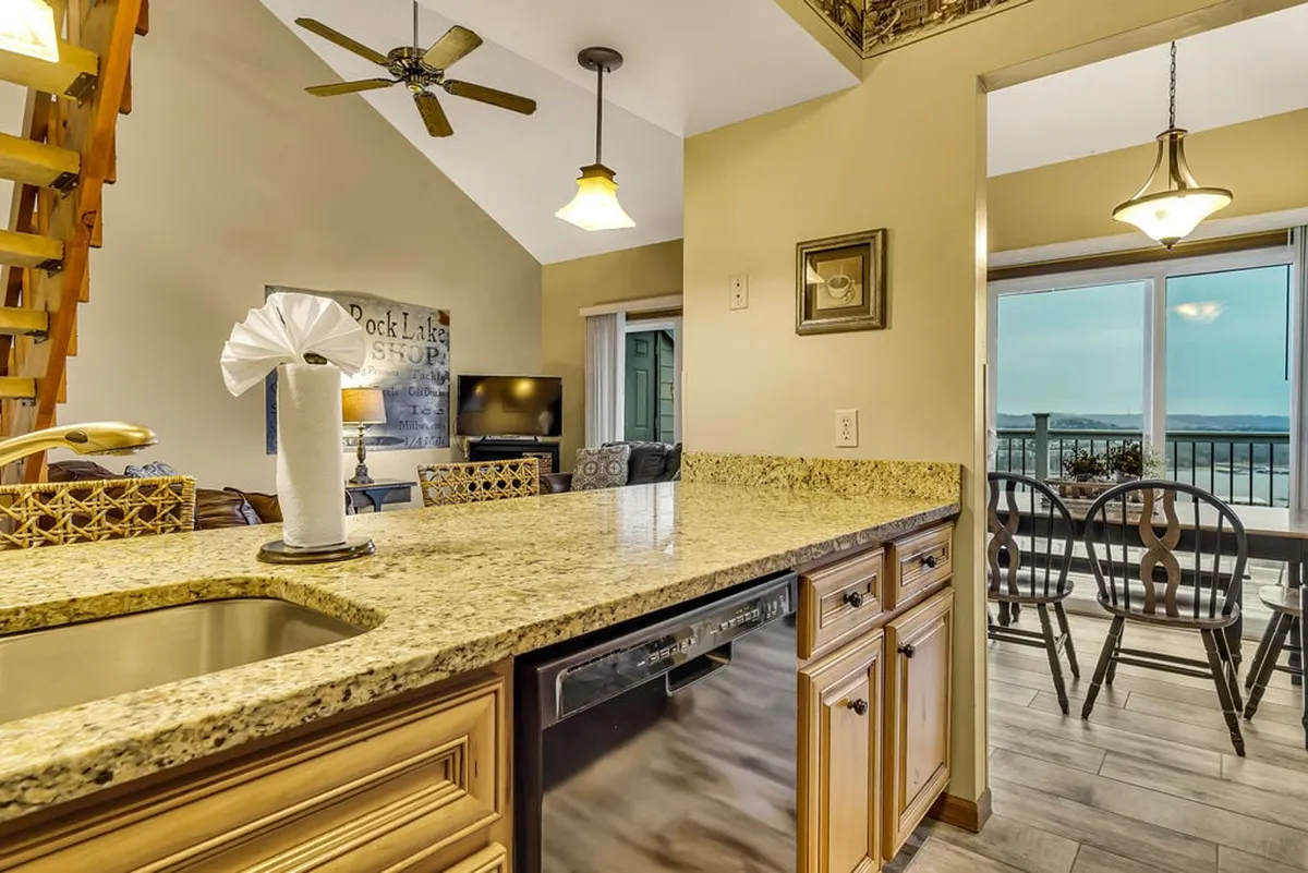 A bright, open-plan kitchen and dining area with granite countertops, modern appliances, and a spacious dining table. Large windows provide a panoramic lake view, flooding the space with natural light.