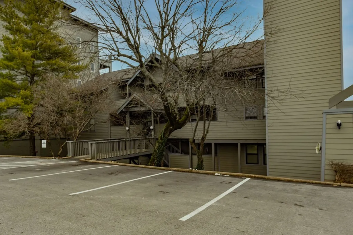 A peaceful exterior view showcasing a wooden walkway that leads past bare trees and toward the condo building’s façade and parking area. The mood is quiet and relaxing, perfect for a morning coffee or an evening stroll.