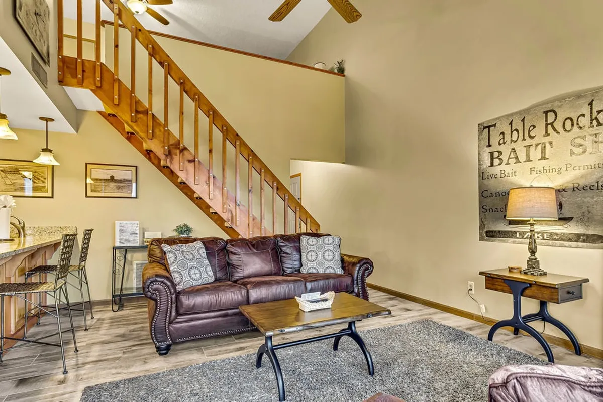 This image reveals the interior living space with a clear view of a wooden staircase leading upwards. The leather sofa, decorative wall art, and bar area create a cozy, lived-in atmosphere, but the staircase is a central visual element.