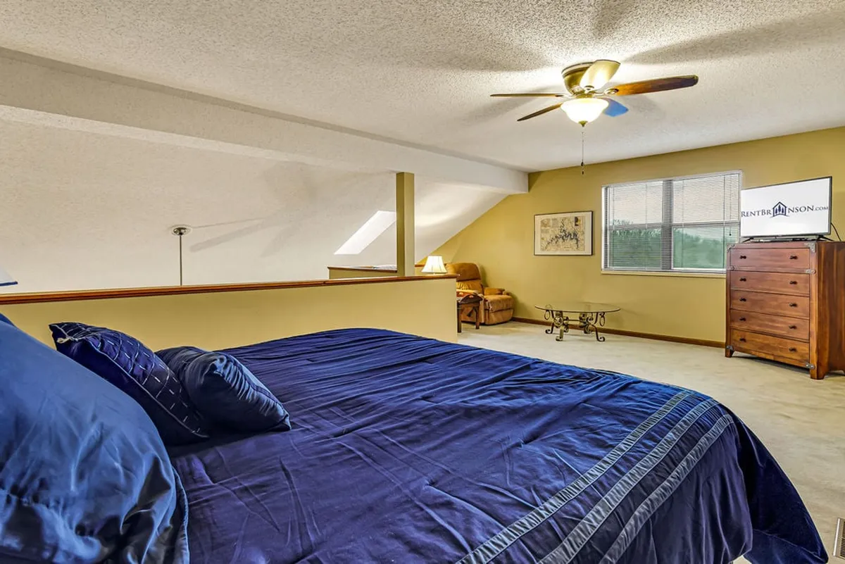 A spacious master bedroom featuring a king-sized bed with elegant navy bedding, a wooden dresser, a comfortable armchair, and a large window letting in natural light. The arrangement is serene, with soft accents and restful decor.